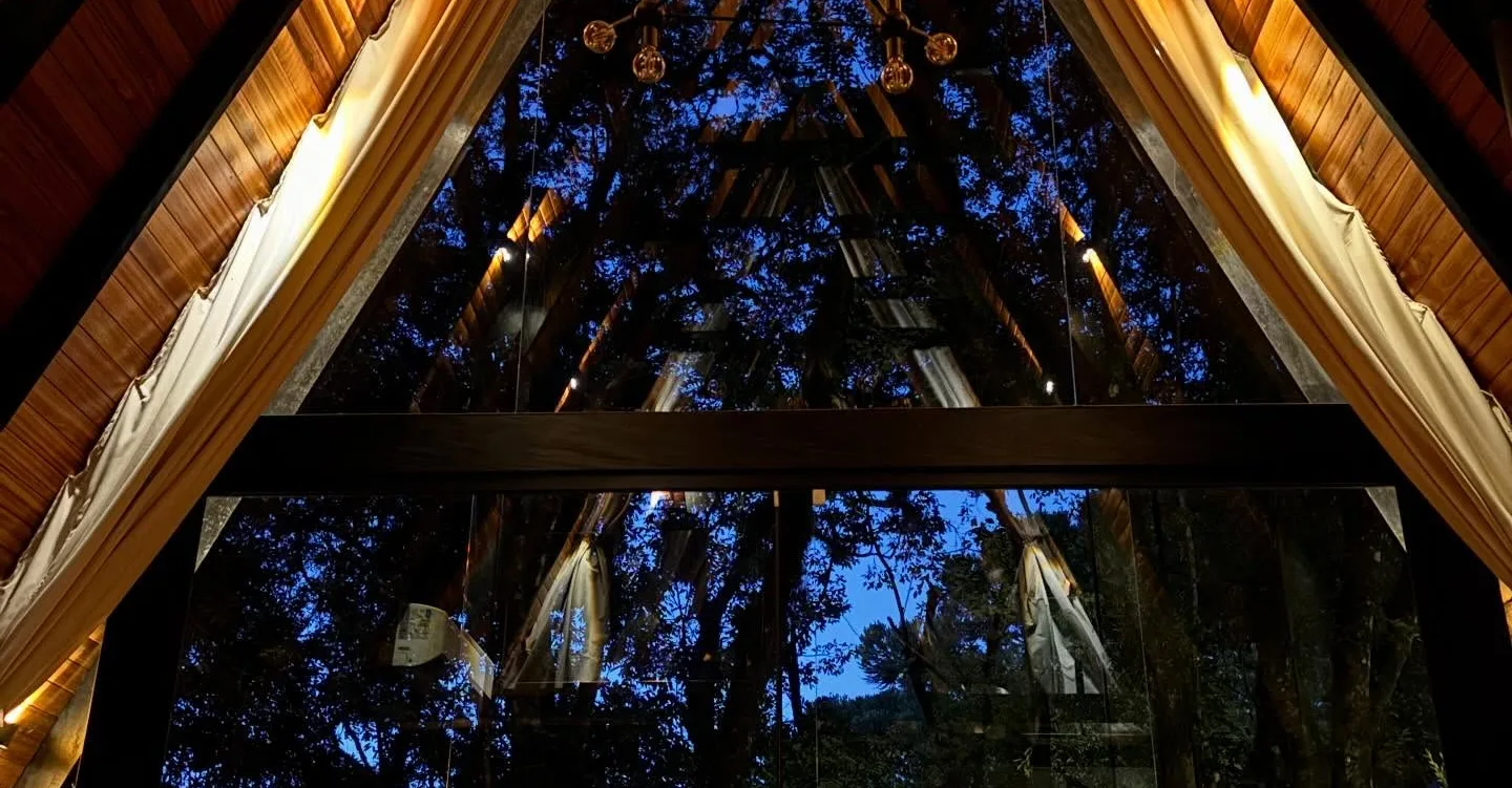 O interior da cabana à noite, sob o telhado em A, com a mata escura pelo vidro